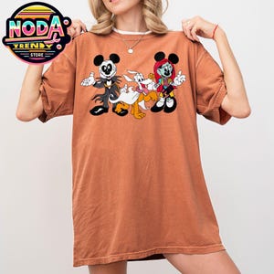May include: An orange-brown t-shirt featuring a cartoon graphic of Jack Skellington, Pluto, and Sally from The Nightmare Before Christmas. The characters are in a Halloween-themed design. The shirt has a crew neck and short sleeves.