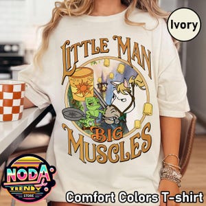 May include: Ivory Comfort Colors T-shirt featuring a cartoon design with the text "Little Man Big Muscles". The graphic includes a horse, a frog, and a sun. The shirt is a light cream color.