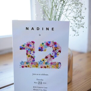 May include: A white invitation card with the text "NADINE is 12" in black font. The number 12 is made of colorful flowers. The invitation includes the date "Thursday Nov. 23 2024", the address "B3L3 Lancers Lane Alesana City, Reis", and the RSVP information "RSVP: Anna 123456789".