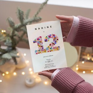 May include: A white invitation card with the name Nadine and the number 12 made of colorful flowers. The text on the card reads "Join us as we celebrate... Thursday, Nov. 23, 2023..."