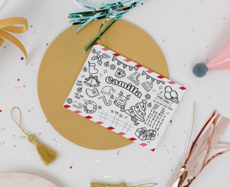 K&ouml;nnte beinhalten: Eine wei&szlig;e Weihnachtskarte mit dem Namen "Camilla" in Schwarz. Die Karte zeigt verschiedene festliche Illustrationen und Wortsuchr&auml;tsel. Sie liegt auf einer goldenen runden Basis, umgeben von dekorativen B&auml;ndern und Konfetti.