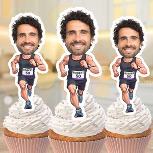 Puede incluir: Tres adornos para cupcakes con una ilustración de dibujos animados de un hombre corriendo con una camiseta sin mangas y pantalones cortos negros. Los cupcakes tienen glaseado blanco y envoltorios rosas. El hombre lleva la inscripción "Robert 50".
