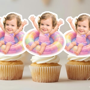 Könnte beinhalten: Drei Cupcakes mit weißem Zuckerguss, jeweils mit einem ausgeschnittenen Bild eines Babys in Badeanzug und Schwimmring. Das Baby trägt einen rosa Badeanzug und lila Shorts. Der Schwimmring ist regenbogenfarben.