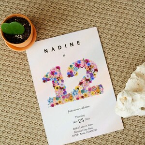 May include: A floral invitation card with the text "NADINE is 12" in a white font on a light pink background. The number 12 is made of colorful flowers. The invitation includes the date "Nov. 23, 2024", the address "B3L3 Lancers Lane, Alesana City, Reis", and the RSVP information "RSVP: Anna 123456789".
