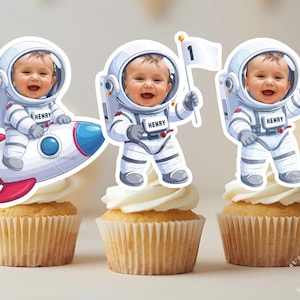 May include: Three cupcakes topped with astronaut-themed decorations. Each decoration features a baby in a spacesuit, with one riding a rocket, one holding a flag, and one waving. The cupcakes have white frosting and are in paper liners.