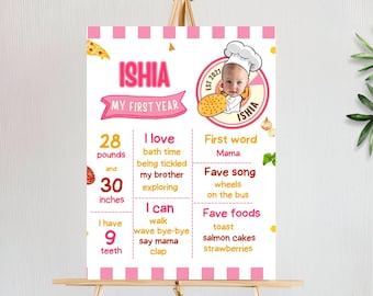 May include: A pink and white milestone poster on an easel. The poster features the name "ISHIA" and "MY FIRST YEAR". It lists the baby's weight as 28 pounds and height as 30 inches. The poster also includes a photo of the baby wearing a chef's hat.