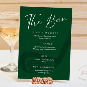 May include: A dark green bar menu with white text listing wines, cocktails, beer, and non-alcoholic beverages. The menu is held up by a cork. Two wine glasses with a golden liquid are in the background.