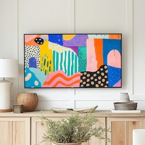Puede incluir: Un salón con un mueble de madera clara y una pintura abstracta colorida. Una lámpara blanca, una cesta de mimbre y objetos decorativos están sobre el mueble. Una mesa de centro con una planta y un sofá blanco completan la escena.