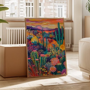 May include: A framed art print depicting a colourful desert scene with cacti, flowers, and mountains under a sunset. The artwork uses blues, oranges, pinks, and greens, creating a lively and eye-catching scene. The frame is a light brown colour.