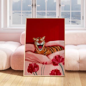 May include: A framed art print featuring a smiling tiger with orange and black stripes, resting on pink pillows and a floral patterned blanket. The background is a solid red, with red flowers in the foreground.