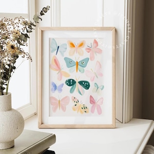 May include: A framed art print featuring a collection of colorful watercolor butterflies. The butterflies are in shades of pink, blue, orange, and green, set against a white background. The frame is made of light-colored wood.