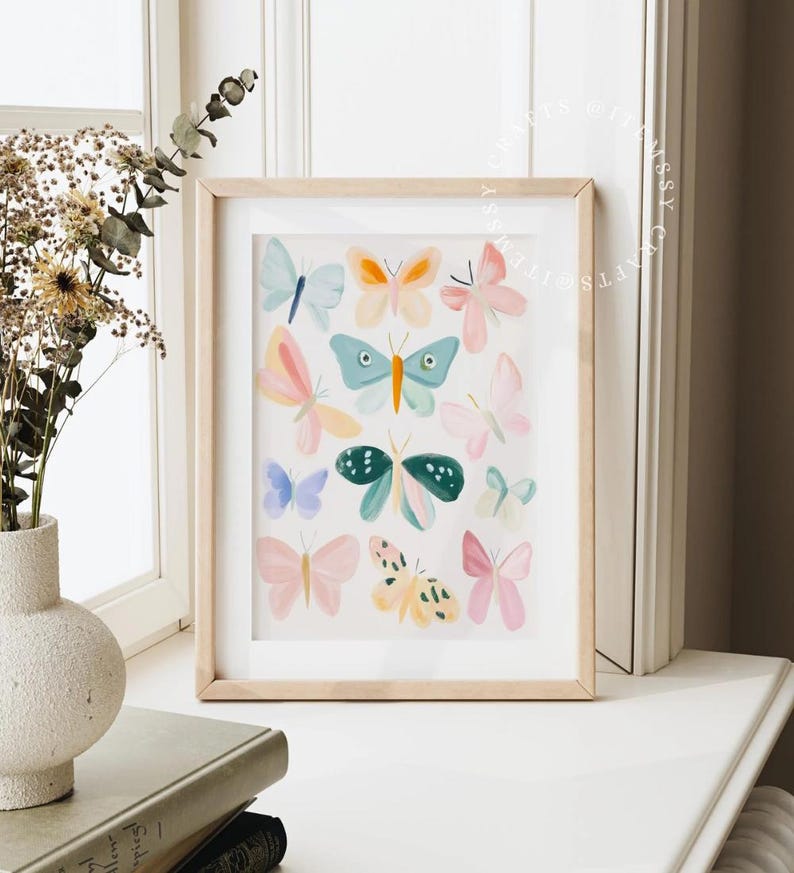 May include: A framed art print featuring a collection of colorful watercolor butterflies. The butterflies are in shades of pink, blue, orange, and green, set against a white background. The frame is made of light-colored wood.