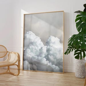 Könnte beinhalten: Ein gerahmter Kunstdruck mit einer Fotografie von flauschigen weißen Wolken vor einem sanften grauen Himmel. Das Kunstwerk wird in einem Raum mit hellem Holzboden, einem Rattanstuhl und einer großen Topfpflanze ausgestellt.