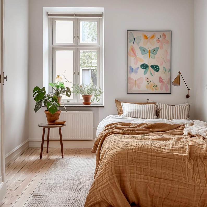 May include: A bedroom with a bed covered in a mustard-colored textured blanket. Two striped pillows and a butterfly art print hang above the bed. A small wooden table holds a potted plant, and a window with plants sits in the background.