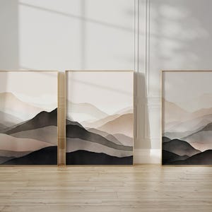 Può includere: Tre stampe artistiche incorniciate di paesaggi. Ogni stampa presenta un dipinto ad acquerello di montagne a strati nei toni del nero, grigio e beige. Le cornici sono color oro chiaro e le opere d'arte sono esposte su una parete chiara.
