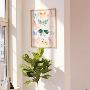 May include: A framed art print featuring a collection of colorful butterfly illustrations. The butterflies are in various pastel shades of pink, blue, orange, and green. The artwork is displayed on a white wall, with a wooden frame. A green plant is in the foreground.