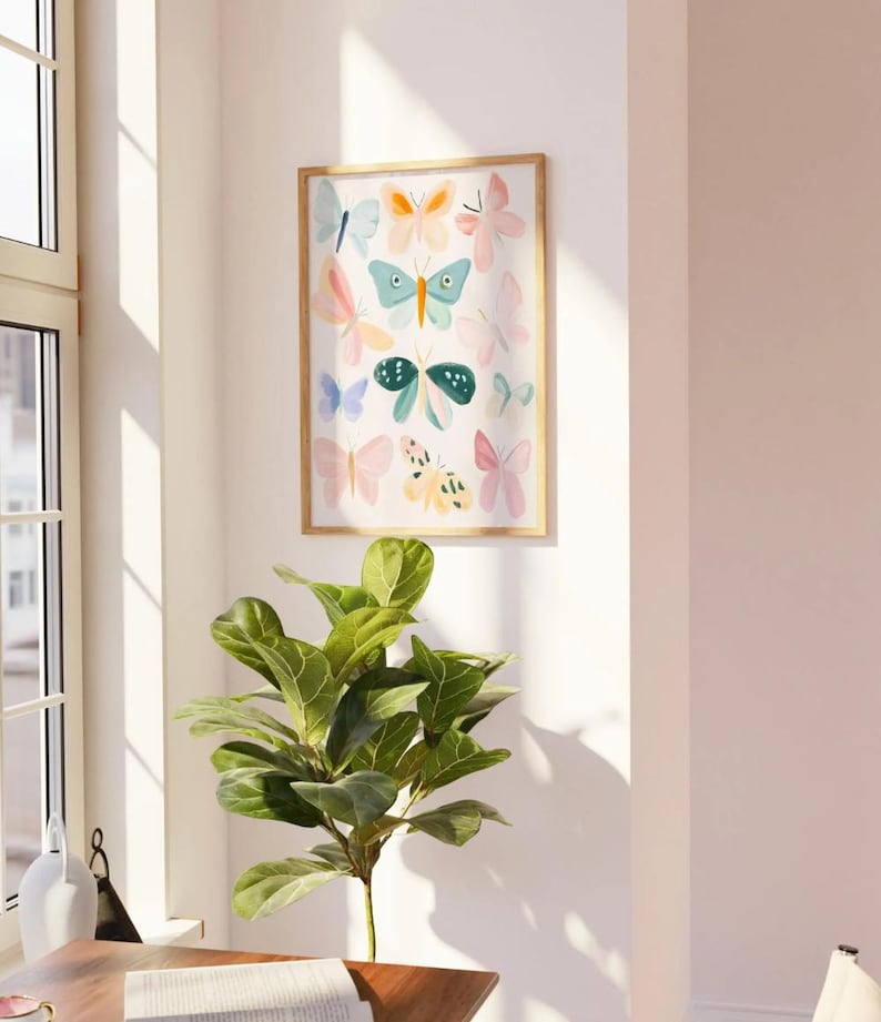 May include: A framed art print featuring a collection of colorful butterfly illustrations. The butterflies are in various pastel shades of pink, blue, orange, and green. The artwork is displayed on a white wall, with a wooden frame. A green plant is in the foreground.