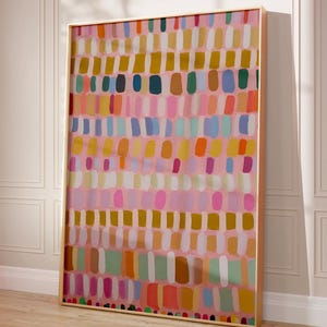 May include: Abstract art print in a gold frame. The artwork features a pink background with rows of colorful, vertical brushstrokes in shades of yellow, blue, orange, green, and white. The framed art is leaning against a white wall and a wooden floor.