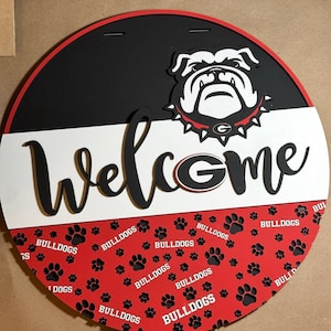 Op de afbeelding: Een ronde houten plaat met een zwart, rood en wit ontwerp. Het bovenste gedeelte is zwart met een bulldoglogo. Het woord "Welcome" staat in grote, zwarte, cursieve letters. Het onderste gedeelte is rood met zwarte pootafdrukken en het woord "Bulldogs".