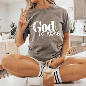 Könnte beinhalten: Ein graues T-Shirt mit dem Text "God is able" in weißen Buchstaben. Der Text ist mittig auf dem Shirt platziert und enthält ein kleines Kreuz über dem Wort "God".