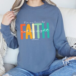 Puede incluir: Una sudadera azul con el texto "For we live by FAITH not by sight 2 Corinthians 7" en un esquema de colores arcoíris.