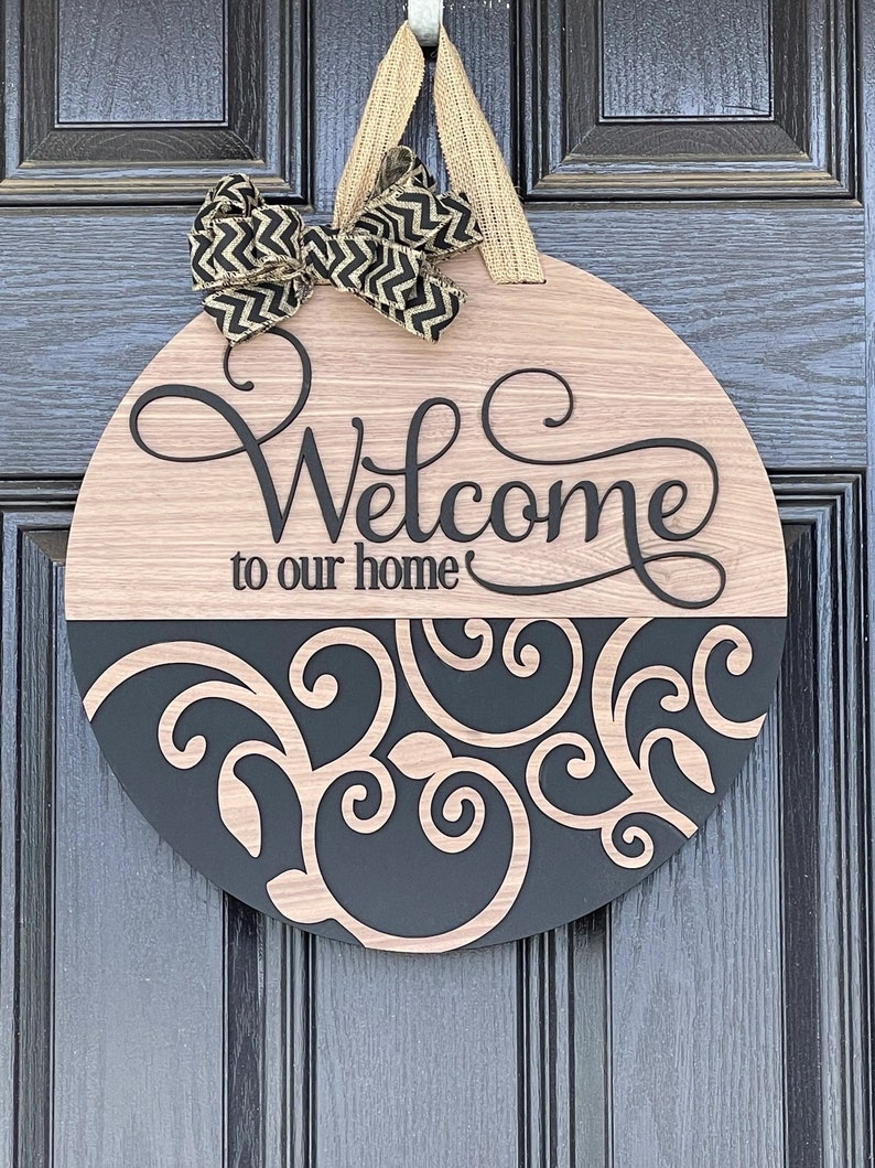 Puede incluir: Un colgador de puerta redondo de madera con un dise&ntilde;o en blanco y negro. La mitad superior del colgador es de madera marr&oacute;n claro con las palabras "Bienvenido a nuestro hogar" en escritura negra. La mitad inferior del colgador es negra con un dise&ntilde;o de remolino decorativo en madera marr&oacute;n claro. Un lazo negro y dorado est&aacute; unido a la parte superior del colgador.