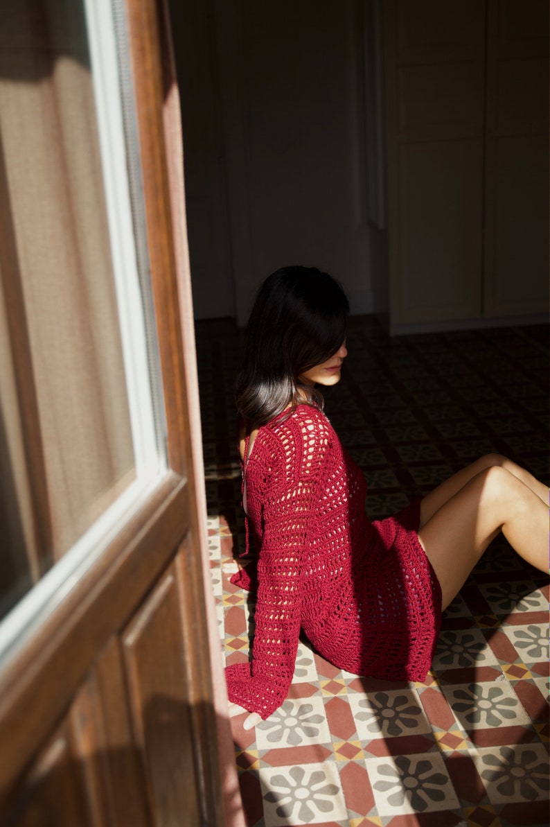 May include: A burgundy crochet sweater dress with long sleeves. The open weave design allows light to pass through. The person is sitting on a patterned tile floor. The dress is a deep red color.