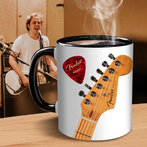 May include: White coffee mug with a black handle and rim, featuring a red Fender guitar pick with "ROAST" text and a guitar headstock design. The mug is on a wooden surface, with a person playing a guitar in the background.
