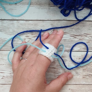 Op de afbeelding: Een hand draagt ​​een witte gehaakte ring met blauw en lichtblauw garen eromheen gewikkeld. Het garen is om de vinger geknoopt. Er zijn twee bollen garen op de achtergrond, een blauwe en een lichtblauwe.