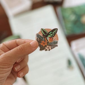 May include: A round sticker featuring a butterfly with green and black wings, orange accents, and a floral design. The sticker is held by a hand, with a blurred background of paper and other stationery items.
