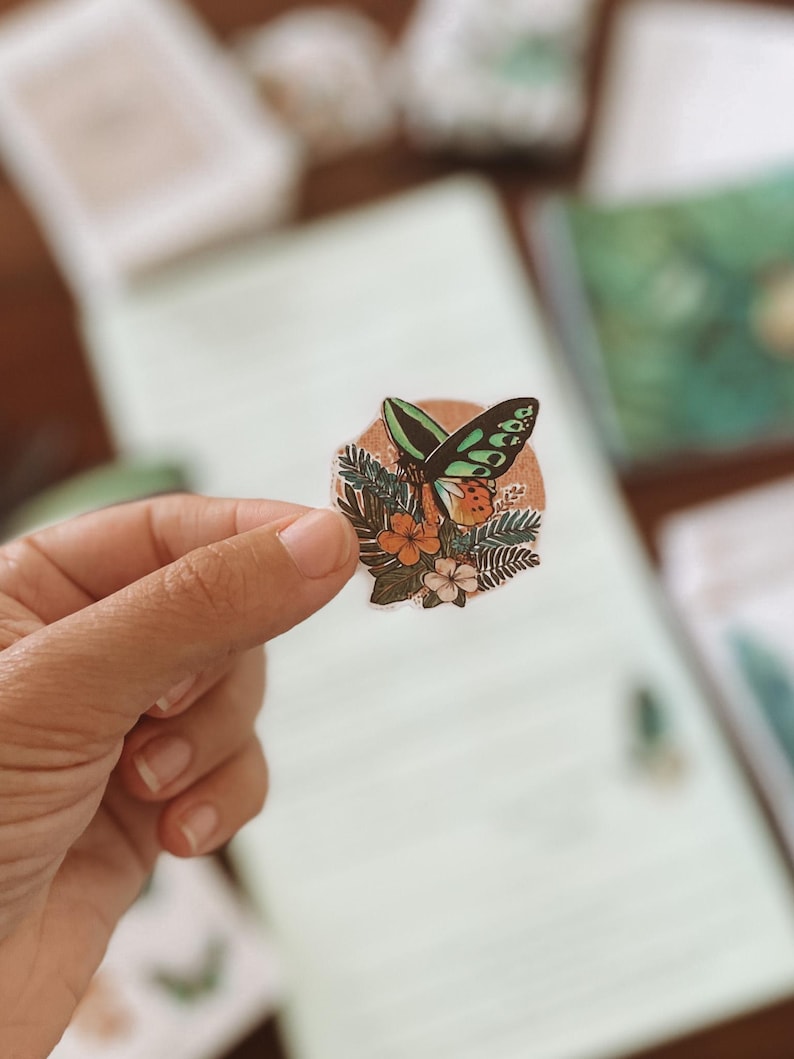 May include: A round sticker featuring a butterfly with green and black wings, orange accents, and a floral design. The sticker is held by a hand, with a blurred background of paper and other stationery items.