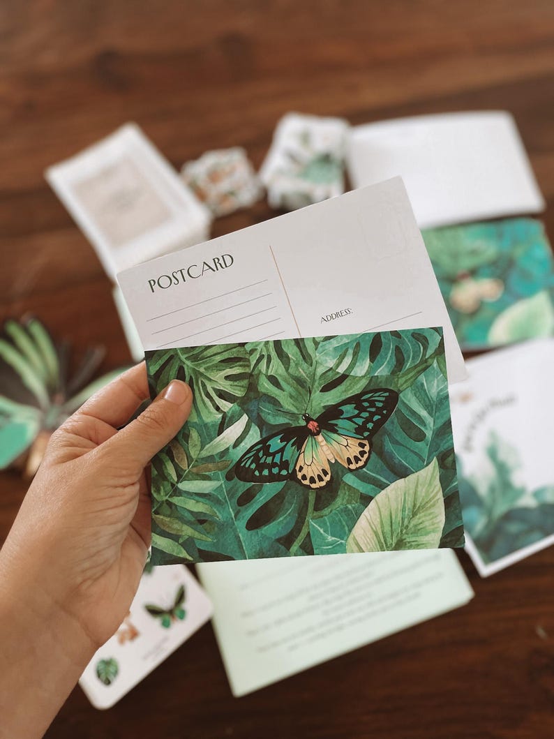 May include: A postcard with a green and black butterfly design, held in a hand. The postcard has the word "POSTCARD" printed on it. The background features other postcards and stationery with similar butterfly and leaf patterns.