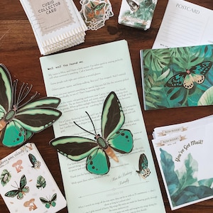 May include: A collection of stationery items featuring butterfly and botanical designs. Includes postcards, stickers, collector cards, and a booklet with text. The color palette consists of green, black, and white, with a wooden background.