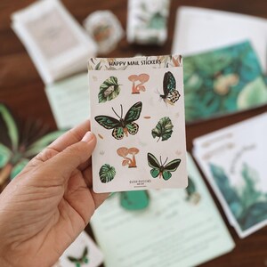 May include: A hand holds a sticker sheet with the text "HAPPY MAIL STICKERS." The stickers feature illustrations of butterflies, mushrooms, and leaves in shades of green and brown. Other stationery items are visible in the background.