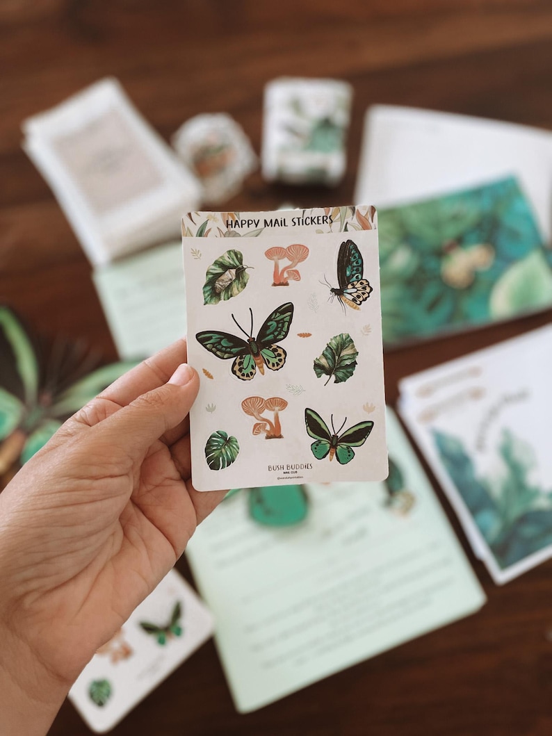 May include: A hand holds a sticker sheet with the text "HAPPY MAIL STICKERS." The stickers feature illustrations of butterflies, mushrooms, and leaves in shades of green and brown. Other stationery items are visible in the background.