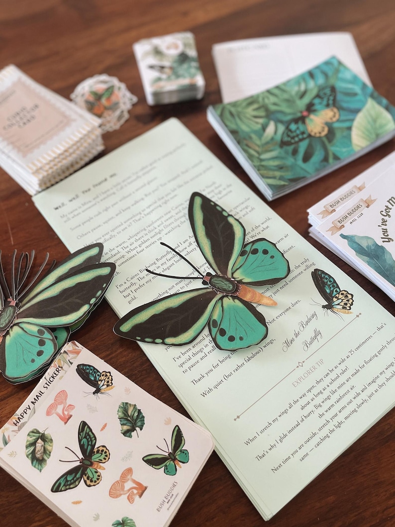 May include: An assortment of stationery items featuring butterfly and botanical designs. Includes stickers, notebooks, and cards with text. The color palette includes teal, black, and green, with a wooden surface.