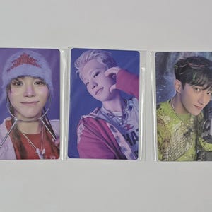 Può includere: Tre fototessere di idoli K-pop in custodie protettive. La prima presenta una persona con un cappello blu e rosso. La seconda mostra una persona con i capelli biondi. La terza mostra una persona che tiene in mano un dispositivo.