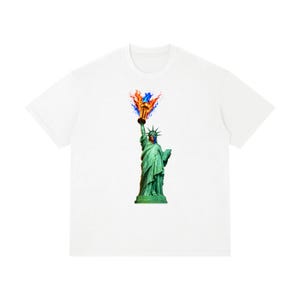 May include: A white short-sleeved t-shirt featuring a graphic print of the Statue of Liberty. The statue is depicted in its characteristic green color, wearing a face mask, and holding a torch with vibrant orange and blue flames rising above.