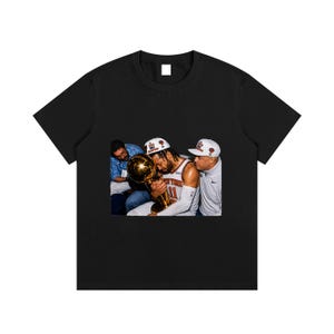 Könnte beinhalten: Ein schwarzes Kurzarm-T-Shirt mit einem zentralen Grafikdruck eines Basketballspielers, der ein weißes Trikot mit orangefarbenem Text und eine weiße Kappe trägt, einen großen goldenen Pokal hält und von zwei Personen flankiert wird.