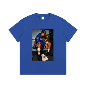 Könnte beinhalten: Ein königsblaues Kurzarm-T-Shirt mit einem rechteckigen Grafikdruck eines Basketballspielers im blau-orangen Trikot, der auf einer Bank sitzt und eine goldene Trophäe hält, mit einem Handtuch auf dem Boden, auf dem NBA CHAMPIONS 2024 steht.