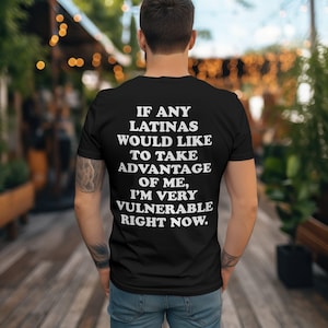 May include: Black t-shirt with white text: "IF ANY LATINAS WOULD LIKE TO TAKE ADVANTAGE OF ME, I'M VERY VULNERABLE RIGHT NOW." The shirt is worn by a person from the back, with a blurred outdoor background.