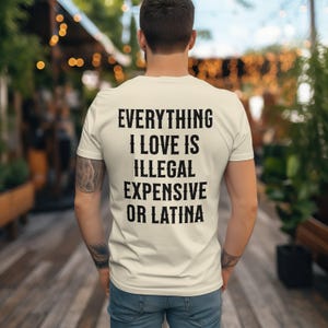 May include: Cream-colored t-shirt with the text "EVERYTHING I LOVE IS ILLEGAL EXPENSIVE OR LATINA" printed in black. The shirt is being worn by a person, viewed from the back, in an outdoor setting.