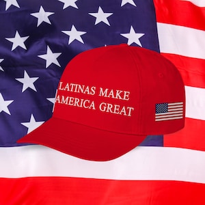 May include: A red baseball cap with the text "LATINAS MAKE AMERICA GREAT" in white. The cap features a small American flag on the side and is set against a backdrop of the American flag.