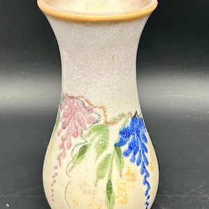 Puede incluir: Un jarrón de cerámica color crema con un acabado moteado. El jarrón presenta un diseño floral con flores rosas, azules y amarillas, junto con hojas verdes. El borde es de color marrón claro.