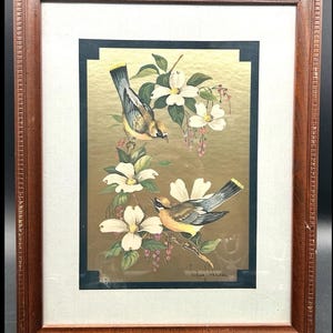 May include: Framed artwork featuring two birds perched on flowering branches. The birds have yellow, gray, and brown feathers. White flowers and pink berries are also depicted against a gold background. The frame is made of brown wood.