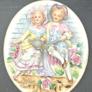 Könnte beinhalten: Eine antike Wandplatte aus Porzellan mit einer 3D-Reliefdarstellung eines jungen Paares in Kleidung aus dem 18. Jahrhundert. Die Frau hält eine Gießkanne und der Mann einen Stock. Sie sind von rosa Rosen und einer Backsteinmauer umgeben.