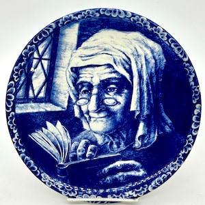 Puede incluir: Plato decorativo azul y blanco con un retrato detallado de una persona mayor leyendo un libro. El plato tiene un borde floral y una ventana en el fondo. La imagen está en una combinación de colores azul y blanco.