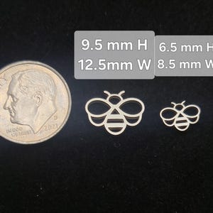 May include: Two silver-colored bee-shaped charms, one larger than the other, next to a US dime. The larger bee measures 9.5 mm high and 12.5 mm wide, while the smaller bee is 6.5 mm high and 8.5 mm wide.