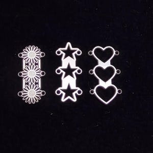 Puede incluir: Tres adornos decorativos de metal. El primero presenta tres flores tipo margarita, el segundo tres estrellas y el tercero tres corazones. Cada diseño tiene pequeños agujeros para fijar.