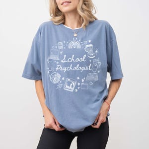 May include: A blue short-sleeved t-shirt with the words "School Psychologist" in white script, surrounded by a circular design of a laptop, coffee cup, rainbow, and other graphics. The shirt is made of a soft material.
